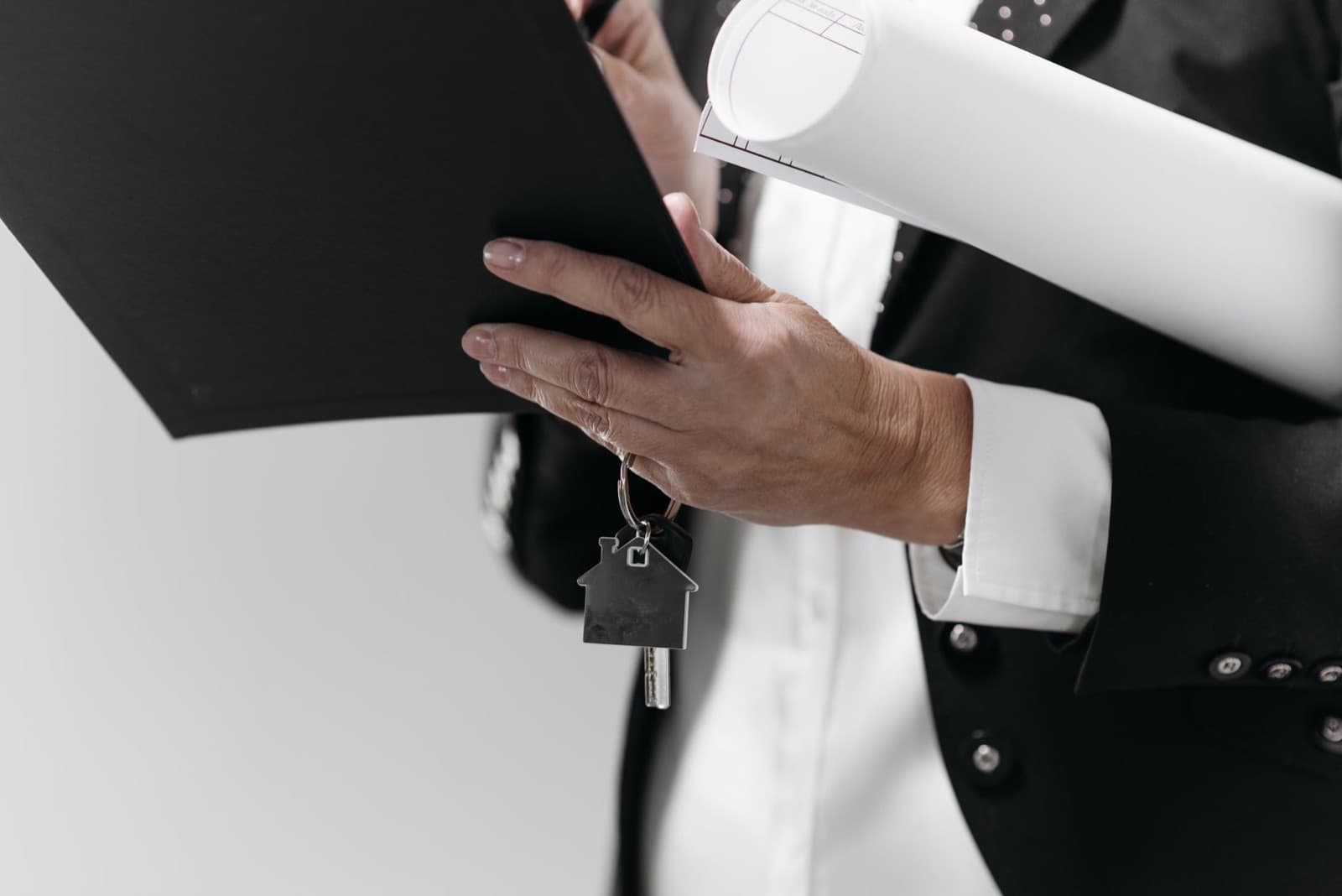 Professional in a suit holding a house keychain, a document folder, and rolled architectural plans.